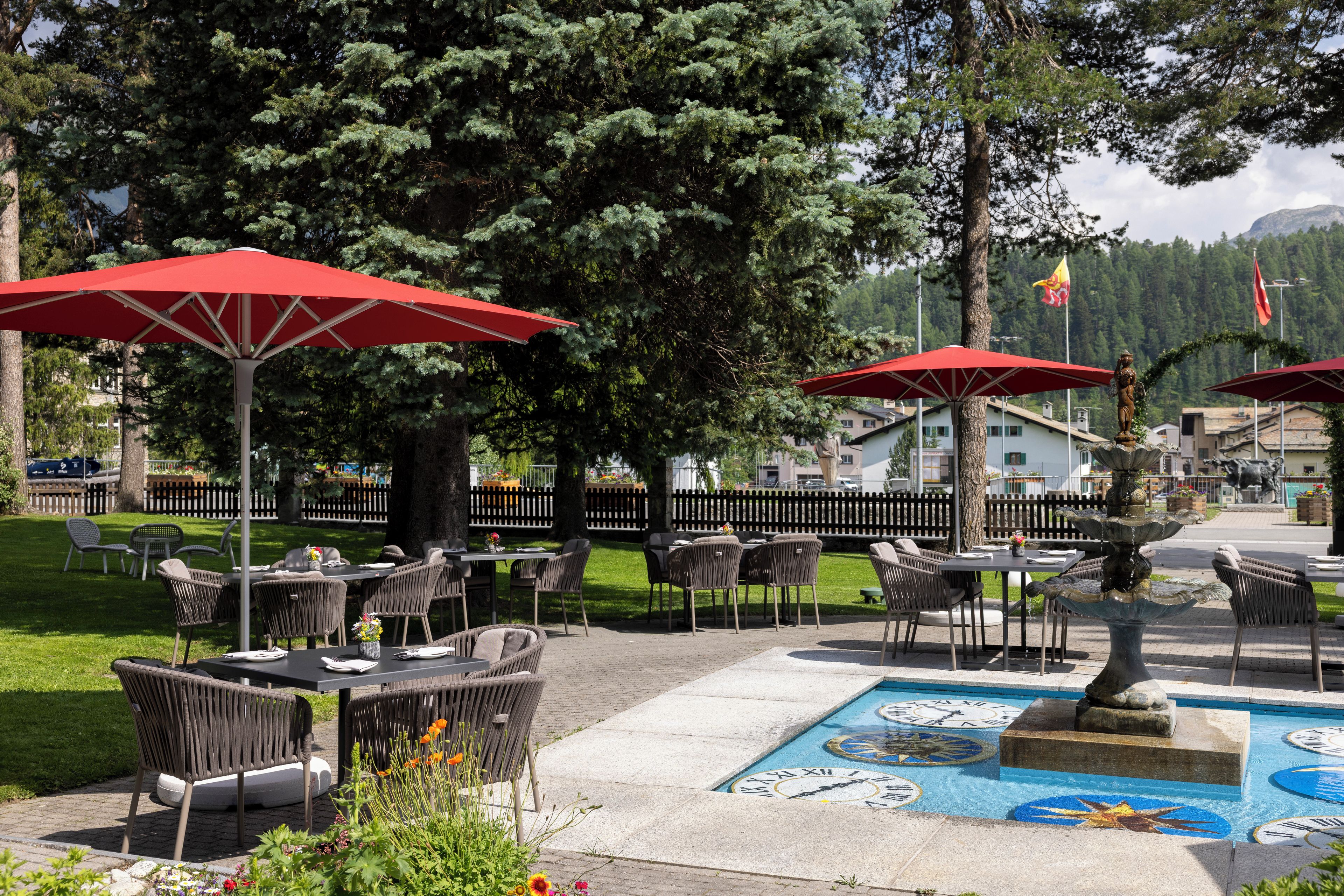 Un espace extérieur aménagé avec des tables et des chaises, des parasols rouges et une fontaine décorative ornée d’une mosaïque représentant une horloge.