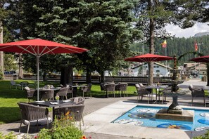 An outdoor seating area with tables and chairs, red umbrellas, and a decorative fountain with a clock mosaic. - Cresta Palace (Celerina-Schlarigna)