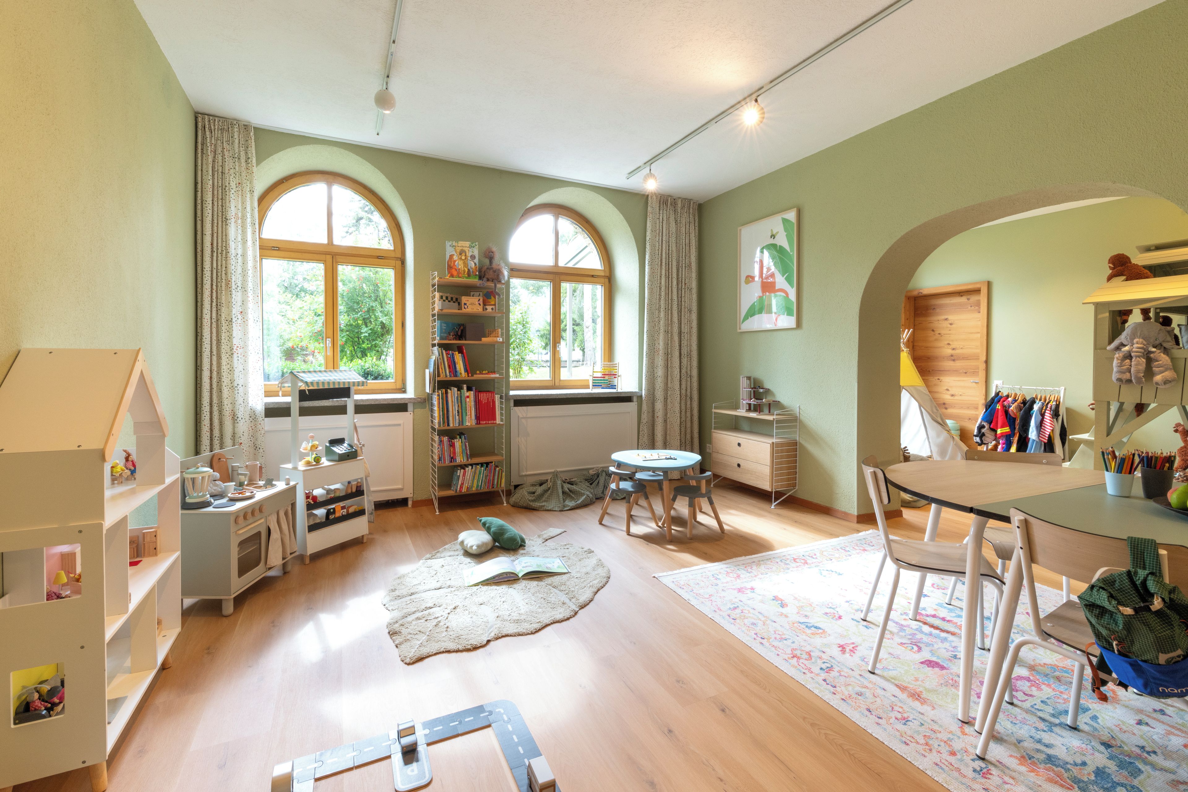 A child's playroom with a wooden floor, a play kitchen, a bookshelf, and a window with a view of trees.