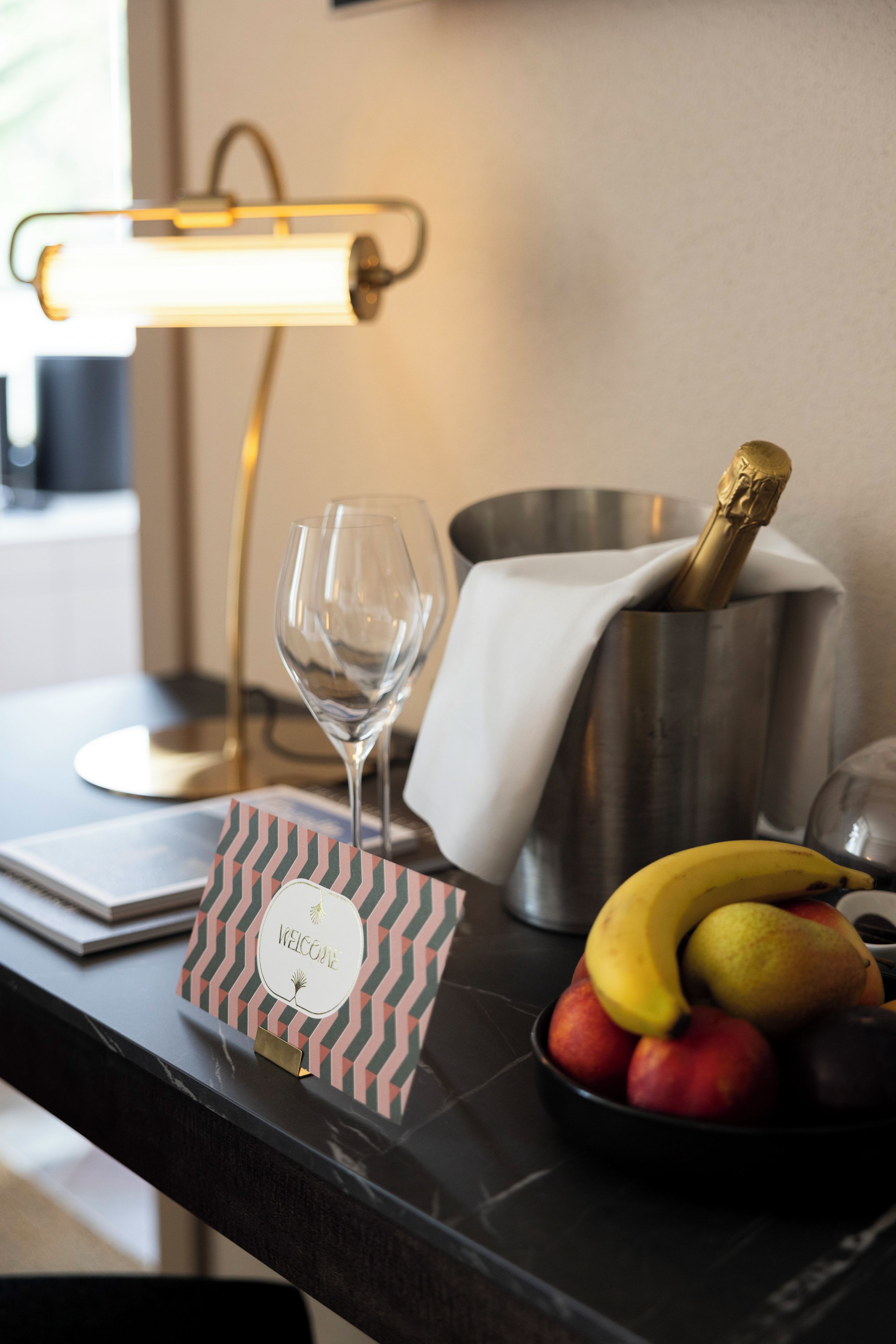 Una mesa con una botella de champán, una copa de vino, un cuenco de fruta y una tarjeta de bienvenida.