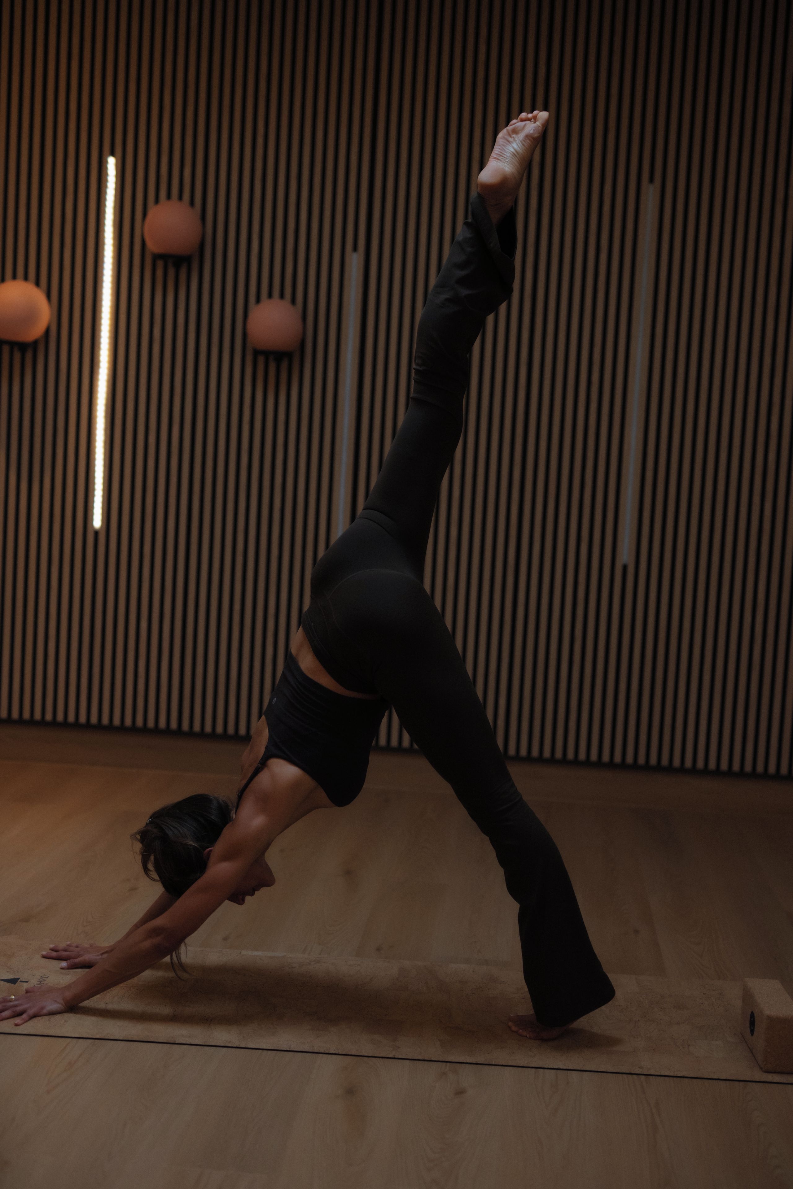 A person performing a yoga pose on a wooden floor with a slatted wall and a wooden cabinet in the background.