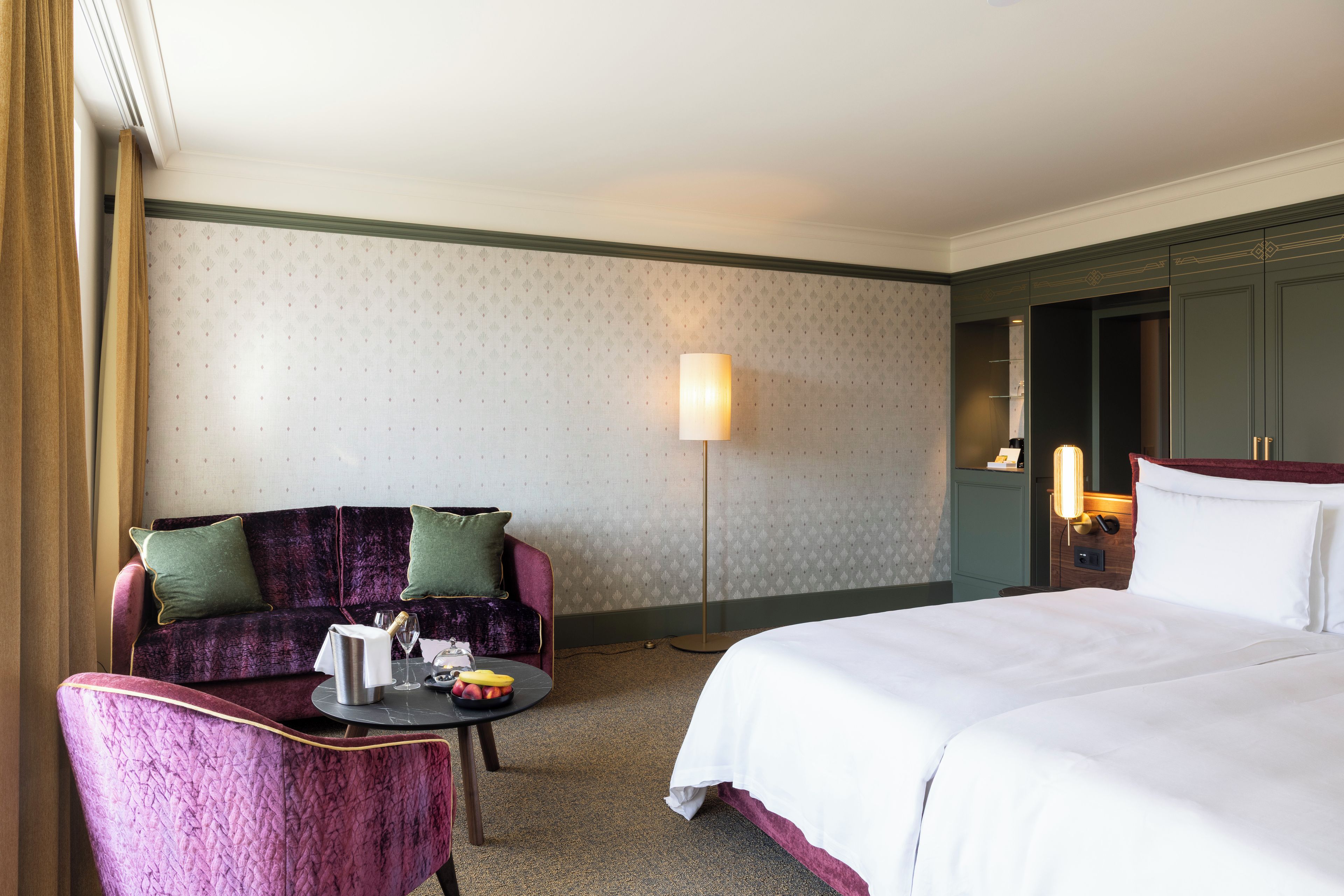Une chambre d’hôtel avec un lit, un canapé, une table sur laquelle se trouve une bouteille de champagne et une lampe.