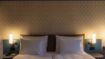 A hotel room with a double bed, two bedside lamps, a nightstand with a phone, and a wall-mounted thermostat.