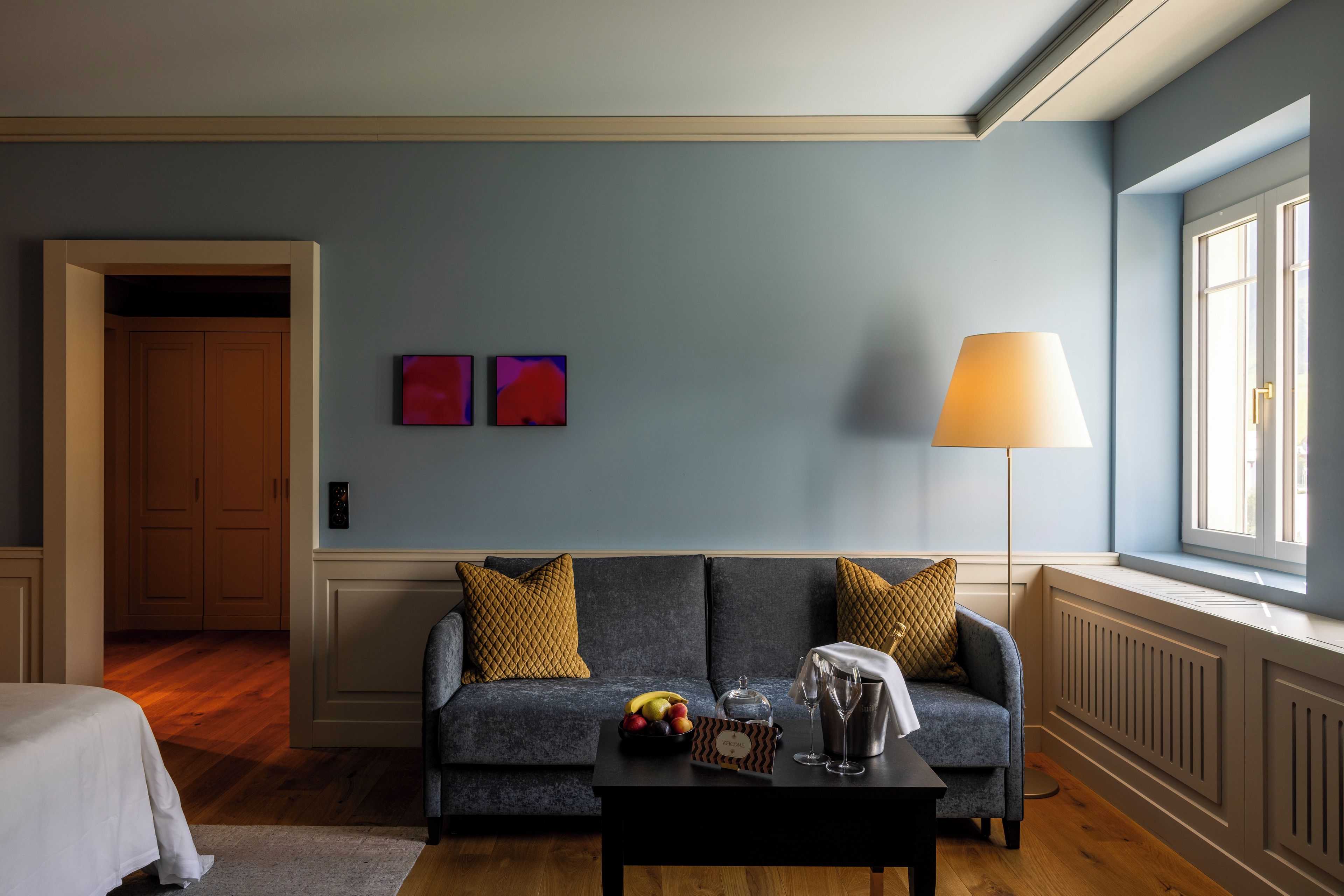 Ein modernes Wohnzimmer mit einem grauen Sofa, einem schwarzen Couchtisch und einer Lampe. An den Wänden hängen zwei Gemälde und ein Fenster bietet einen Ausblick.