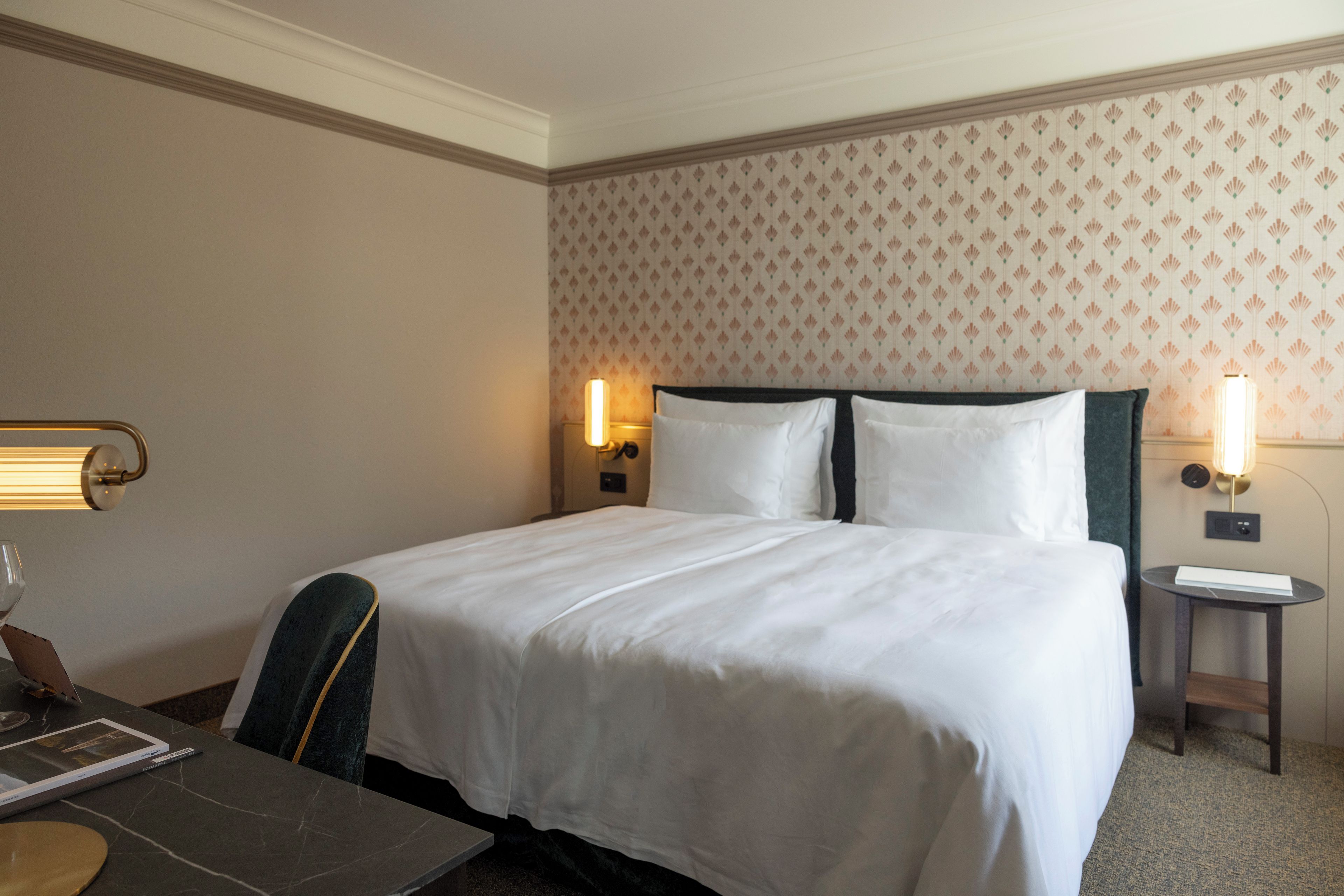 A hotel room with a large bed, two bedside lamps, a small table, and a chair.
