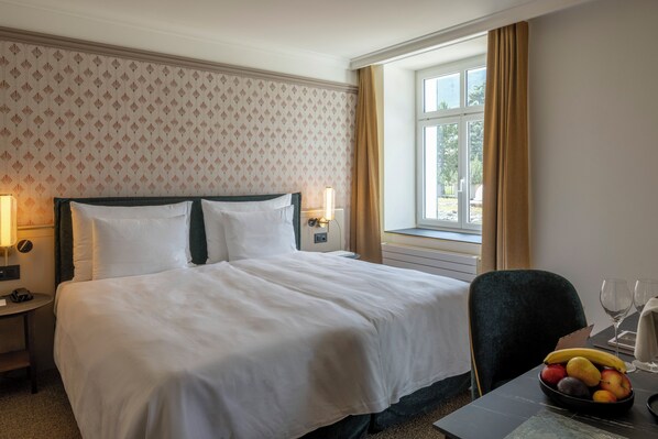 Habitación de hotel con una cama grande, un escritorio con frutero, una silla y un ventanal con cortinas.