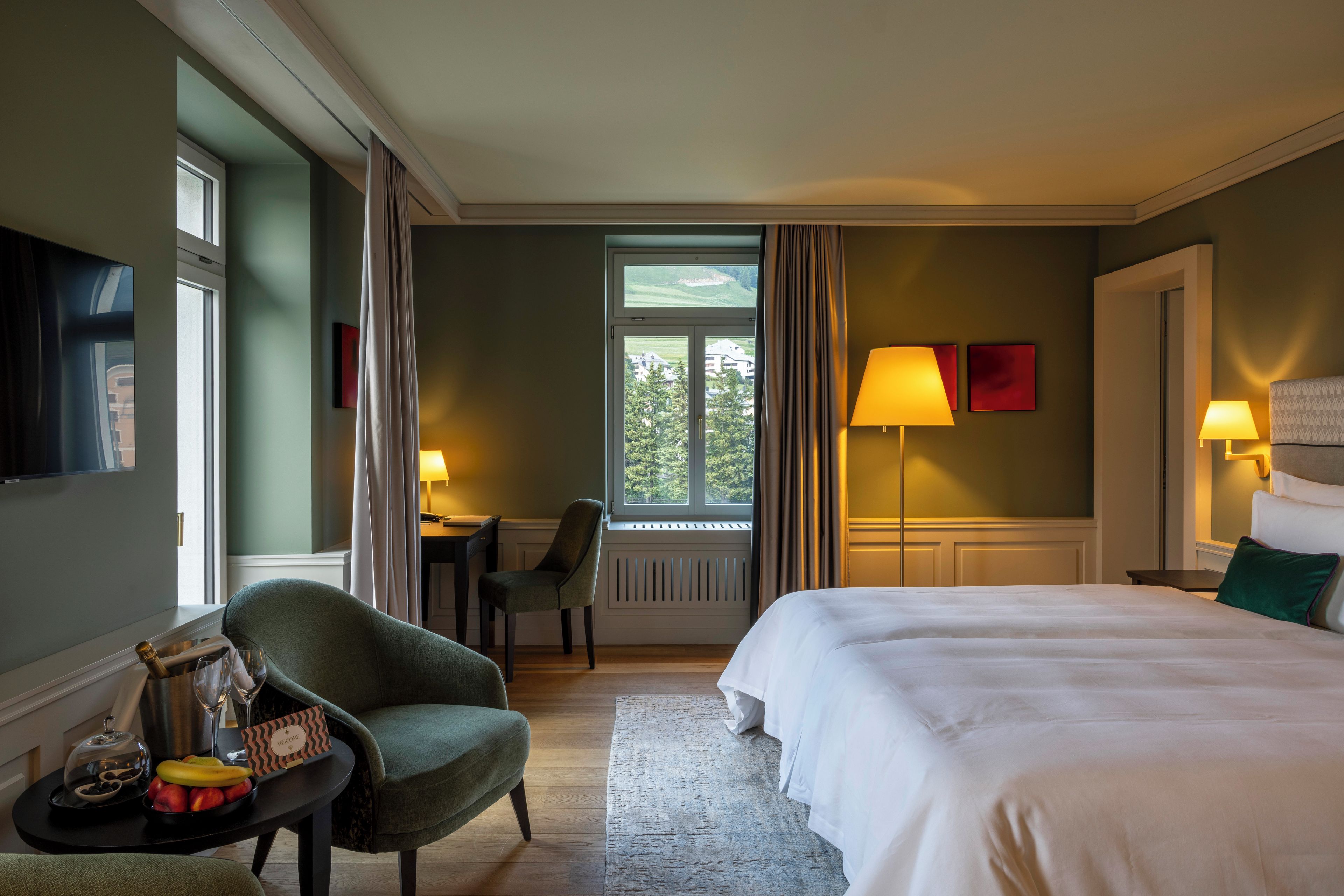 Una habitación de hotel con cama, escritorio, silla, televisor y ventana con cortinas.