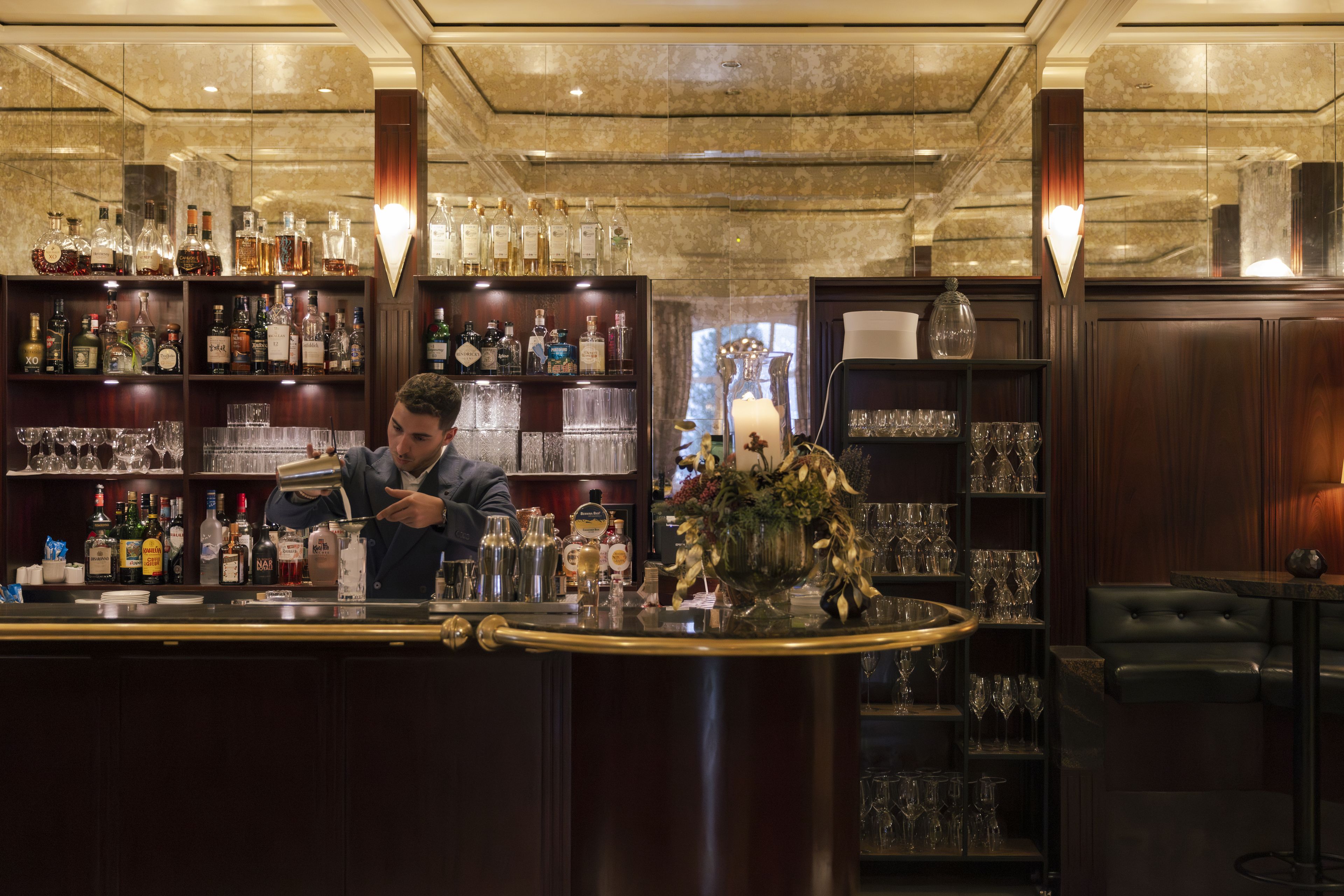 A bar area with a bartender preparing drinks, shelves stocked with various bottles, and a seating area with a vase of flowers.