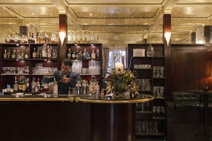 Zona de bar con un cantinero preparando bebidas, estantes llenos de diferentes botellas y un área de asientos con un florero.