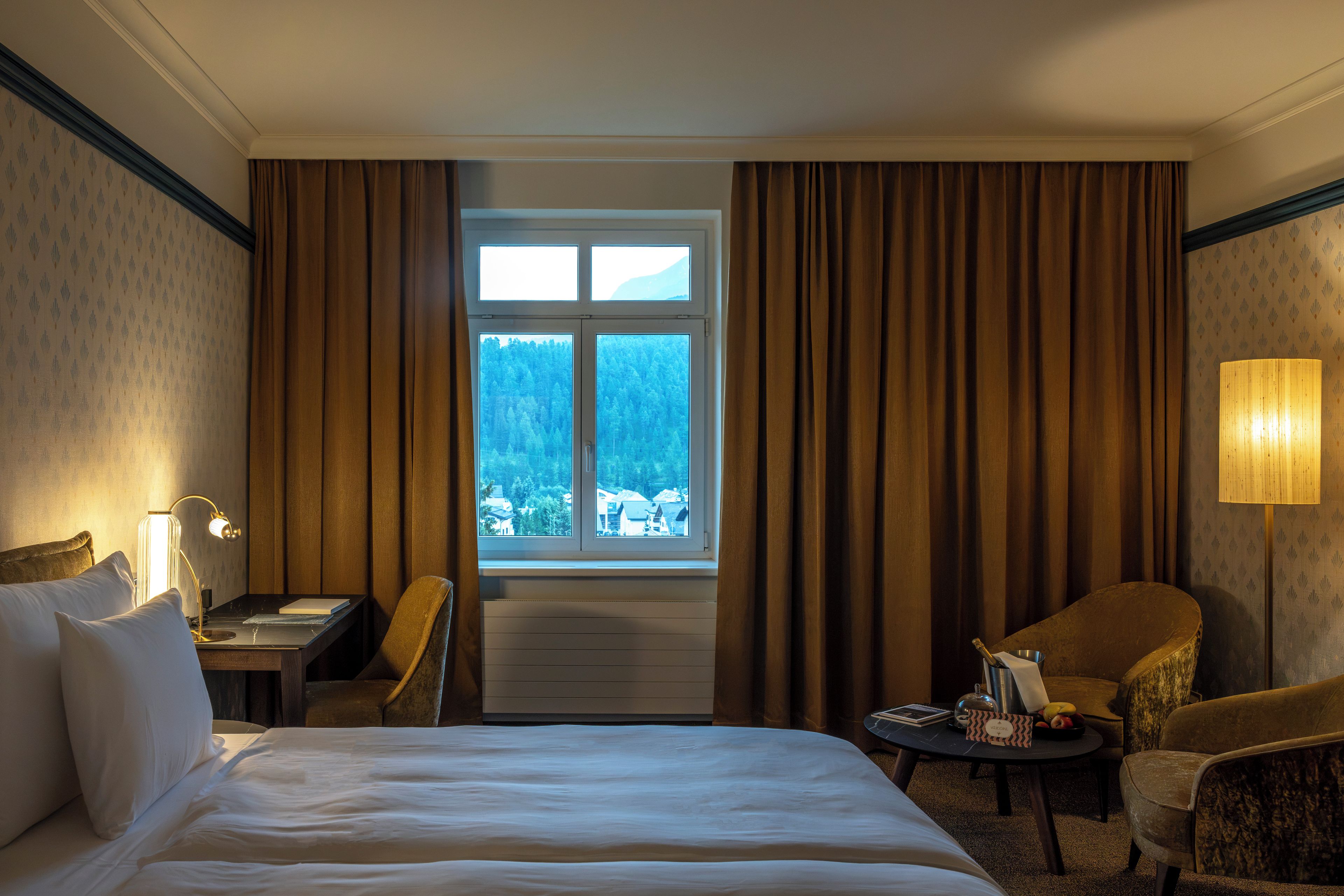 Habitación de hotel con cama, escritorio, silla, mesita de noche con lámpara y vistas a un bosque a través de la ventana.