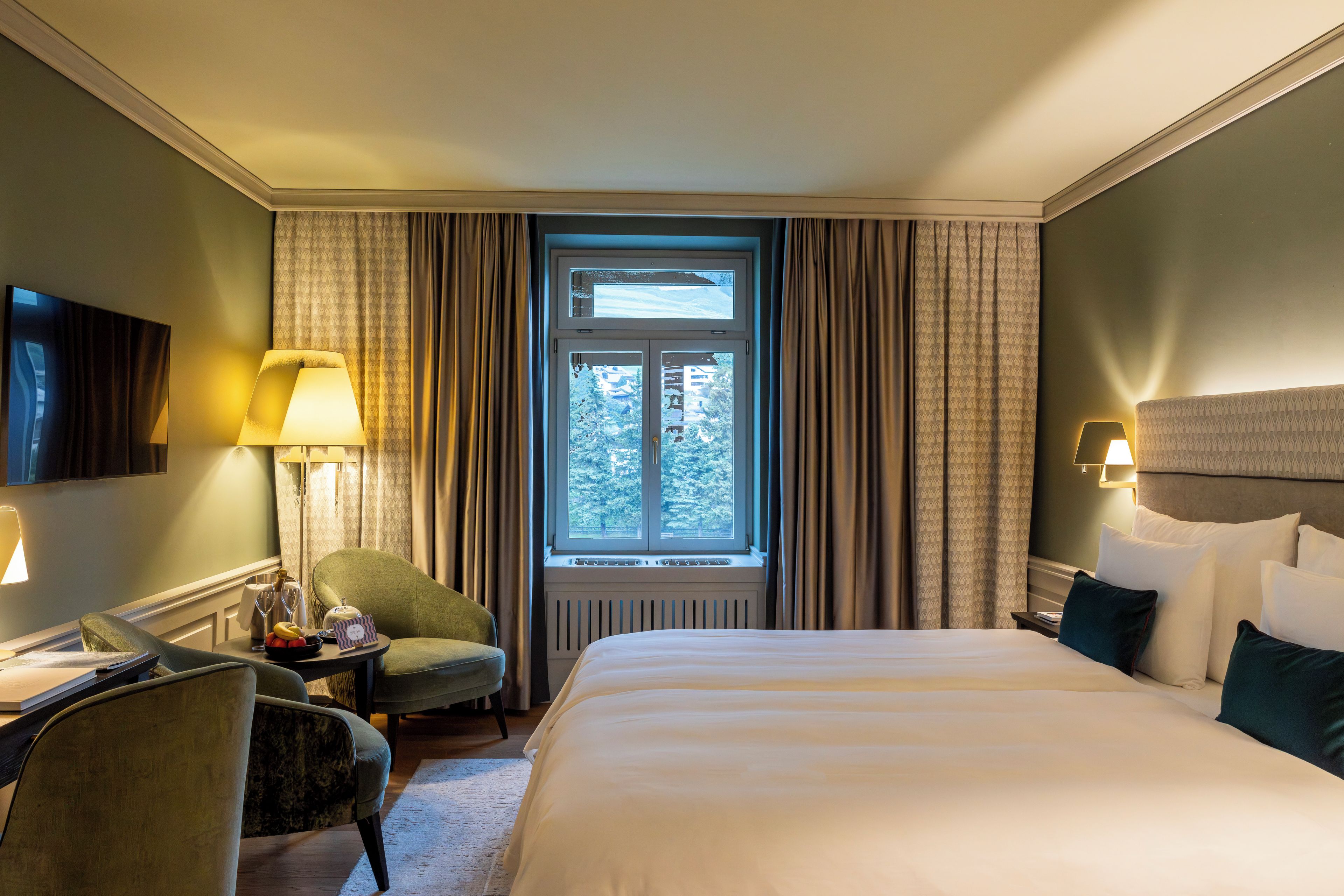 Habitación de hotel con cama, escritorio, silla, ventana con cortinas y televisor en la pared.