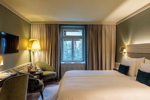 A hotel room with a bed, a desk, a chair, a window with curtains, and a television on the wall.