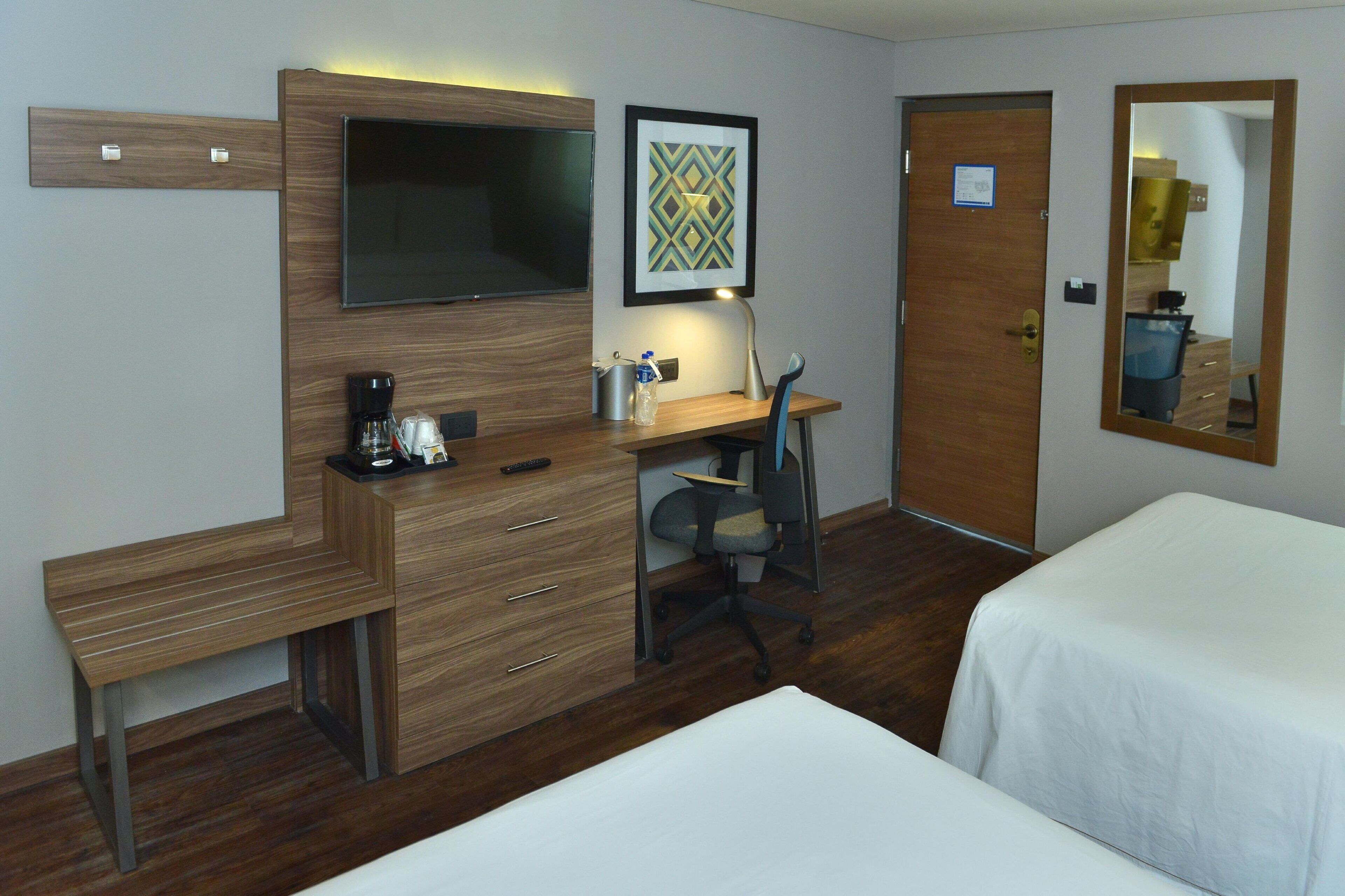 Habitación de hotel con escritorio de madera, televisor de pantalla plana, obra de arte enmarcada, lámpara de escritorio, cafetera, botella de agua, control remoto, una silla y dos camas.