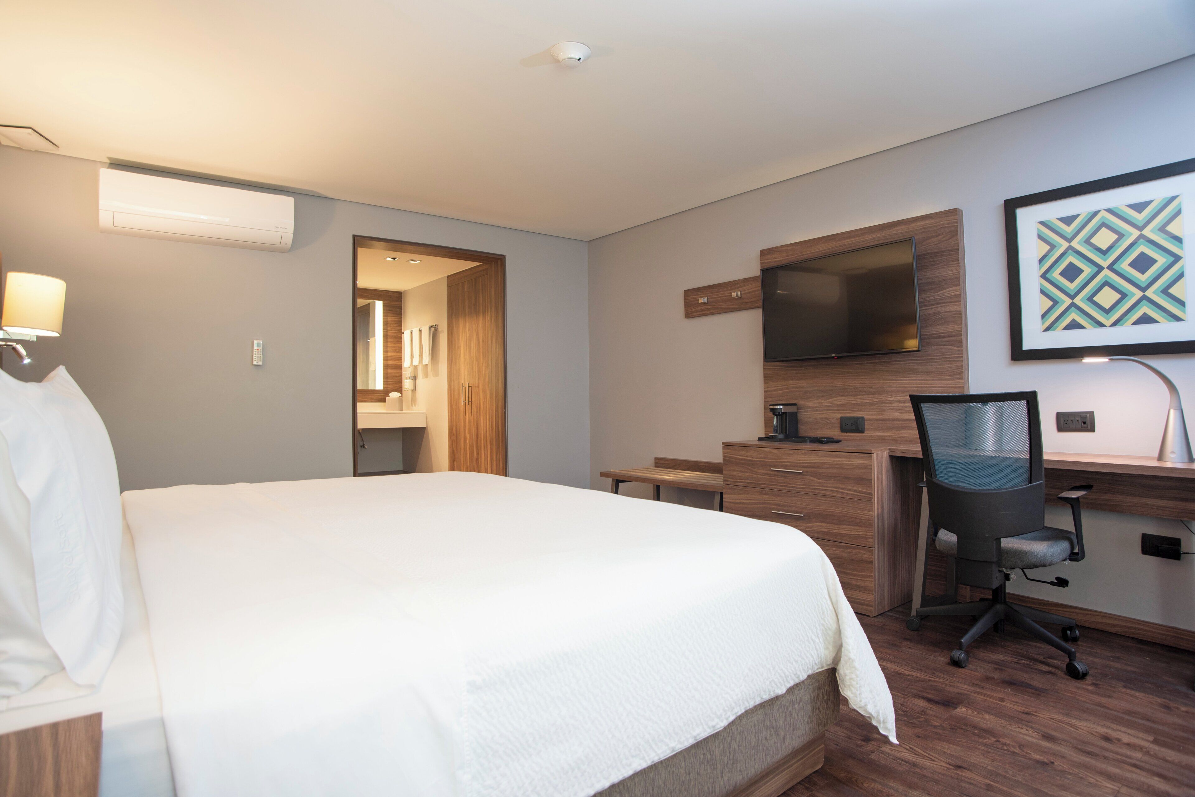 A hotel room with a large bed, a desk with a chair, a TV mounted on the wall, and a framed picture.