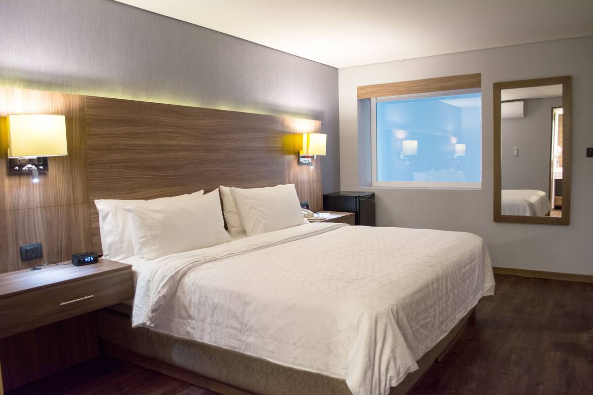 Une chambre d’hôtel avec un grand lit, deux tables de chevet, une commode, une lampe fixée au mur, une fenêtre et un miroir.