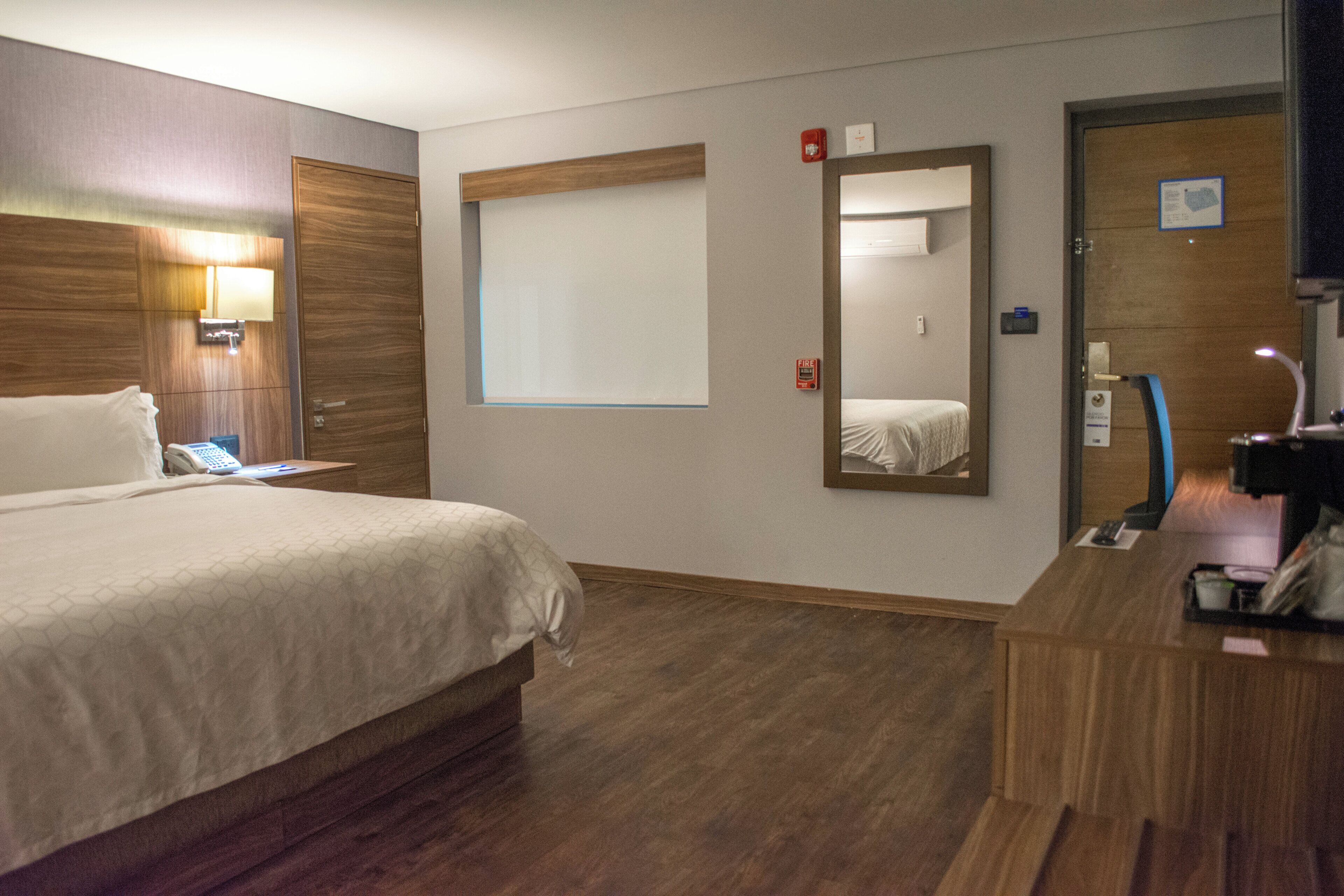 A hotel room with a bed, bedside table, mirror, and a desk with a chair.