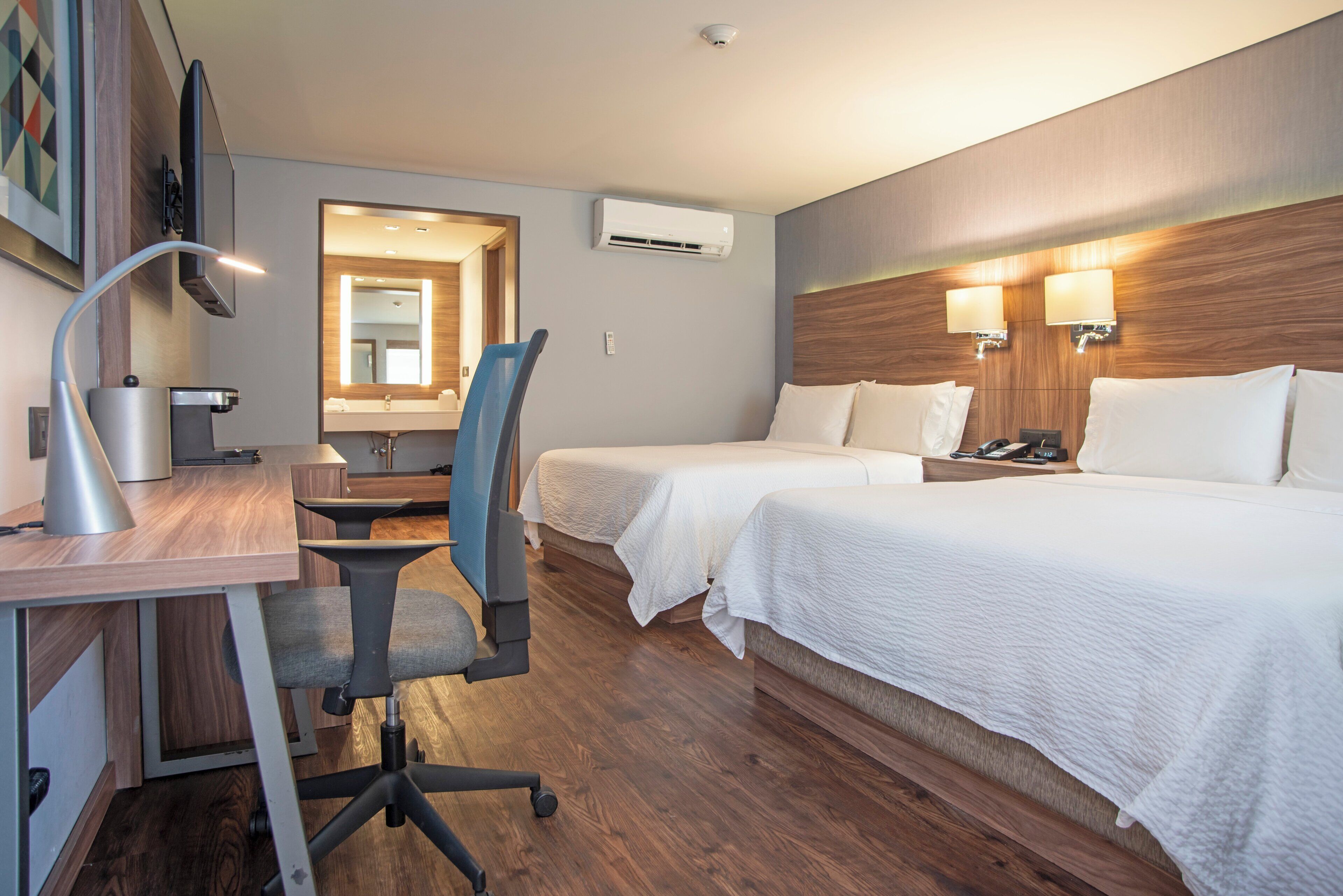 A hotel room with a desk, two beds, a chair, an air conditioning unit, and a mirror.