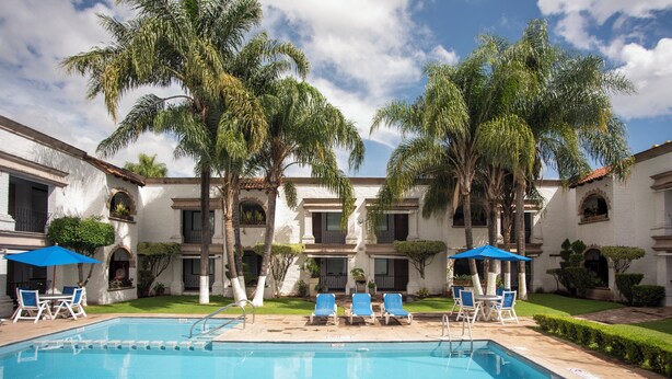 Un área de piscina con sillas y sombrillas azules, rodeada de palmeras y un edificio con techo de tejas.
