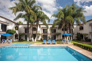 A pool area with blue chairs and umbrellas, surrounded by palm trees and a building with a tiled roof. - Holiday Inn Express - Morelia by IHG (Morelia)