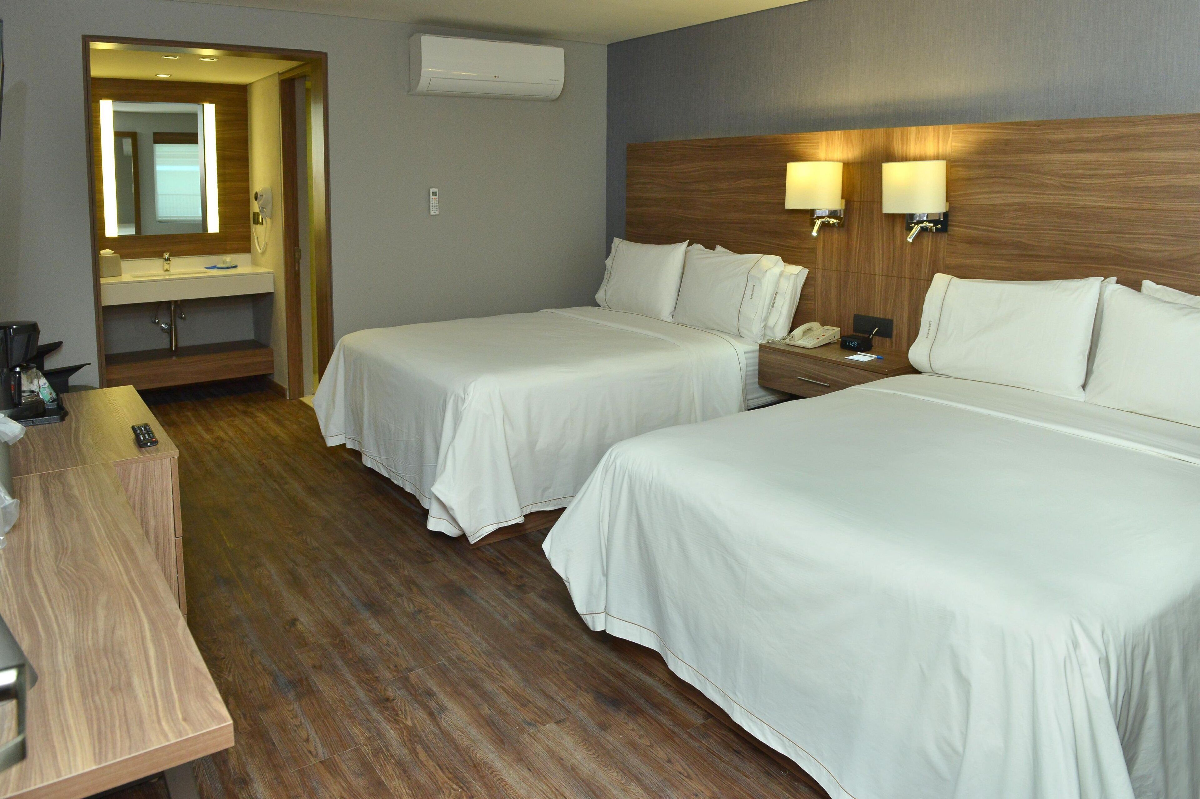 Habitación de hotel con dos camas, un escritorio de madera, un espejo, una mesita de noche con lámpara y aire acondicionado de pared.