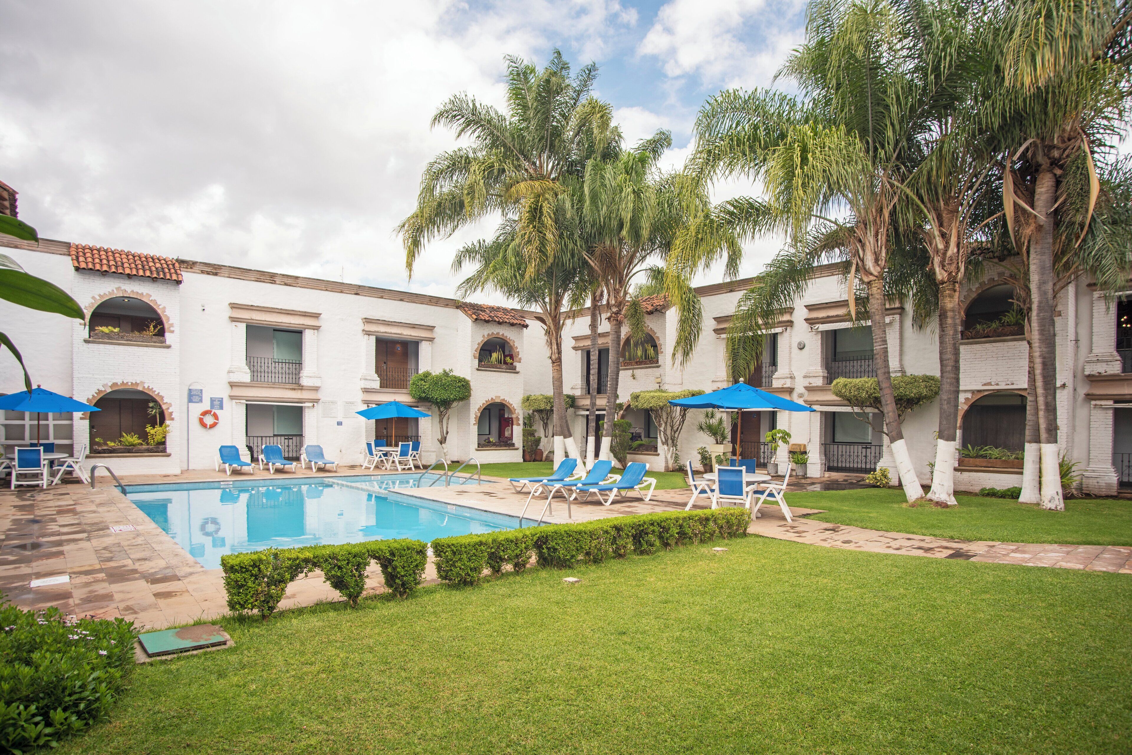 A pool area with lounge chairs and umbrellas, surrounded by palm trees and a building with arched windows.