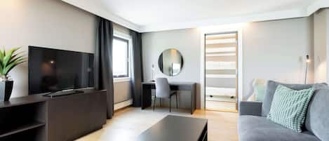 Una sala de estar moderna con un televisor de pantalla plana, un sofá gris, una mesa de centro, un escritorio con una silla y un espejo grande.