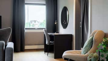 Una sala de estar moderna con un sofá, un escritorio y una silla. Hay una ventana con cortinas, un reloj en la pared y una planta.