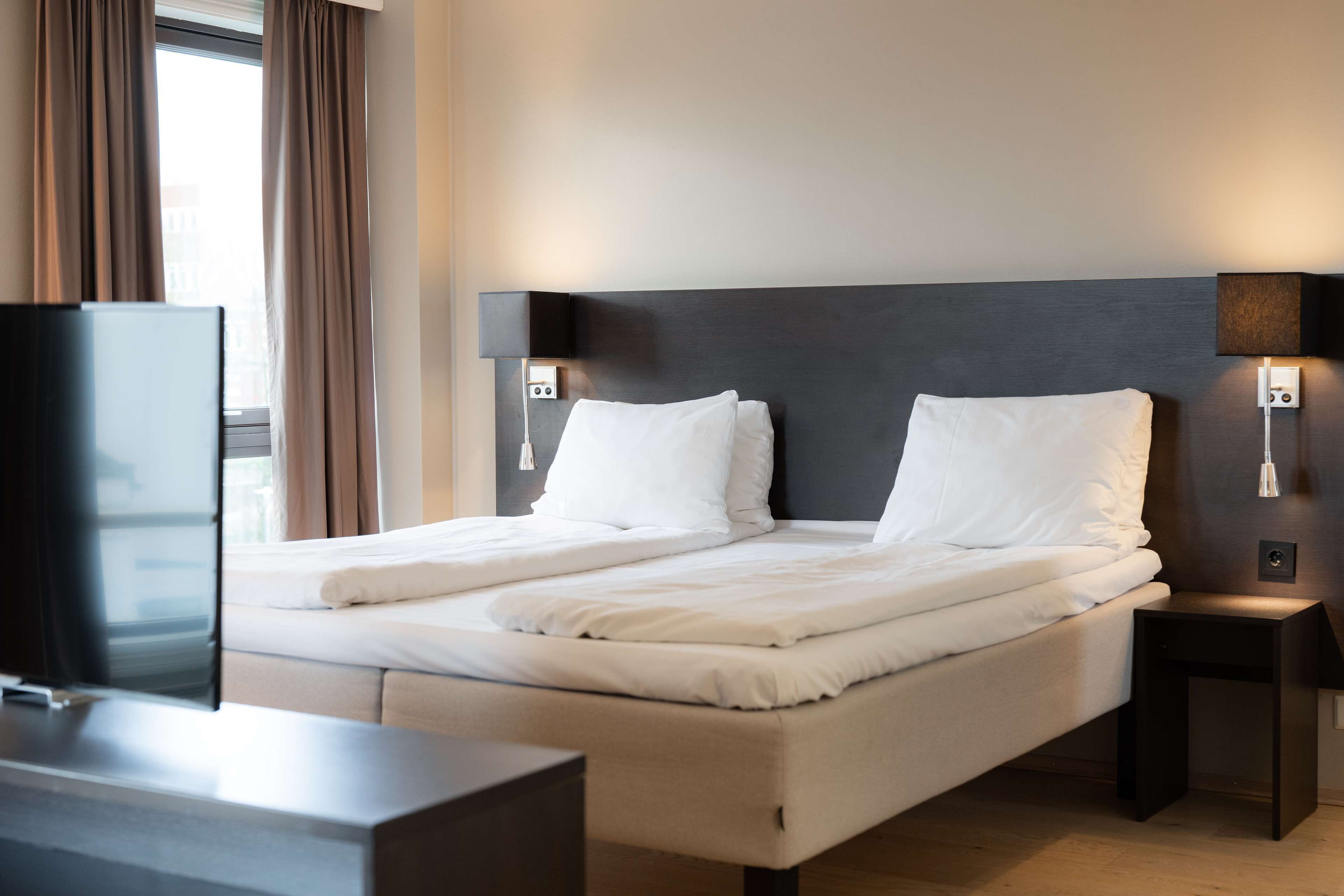 A neatly made double bed with white linens, a dark headboard, bedside lamps, and a flat-screen TV on a stand.