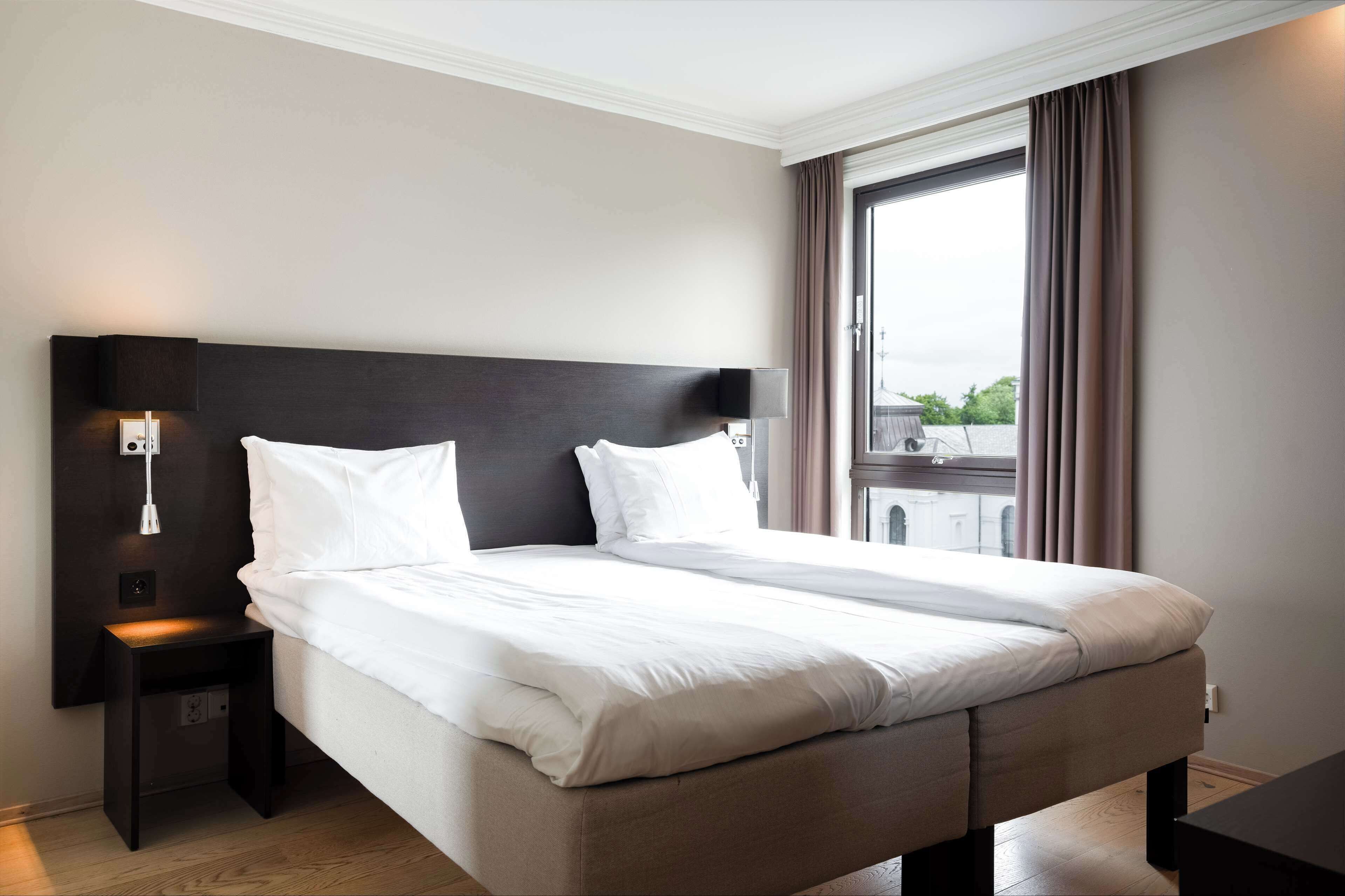 Une chambre d’hôtel moderne avec un grand lit, une table de chevet avec une lampe et une vue sur la ville par la fenêtre.