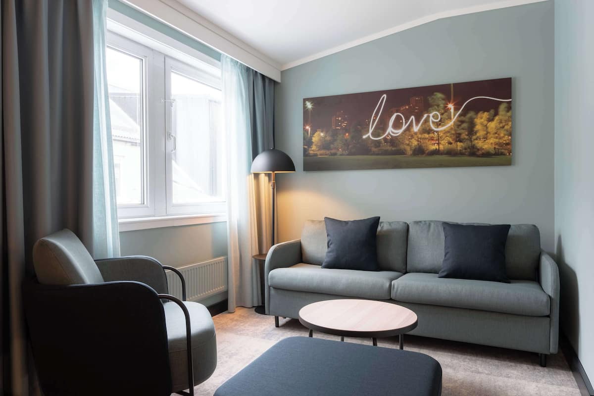 A modern living room with a grey sofa, two armchairs, a small round table, and a floor lamp. A large window with curtains is on the left, and a cityscape painting with the word "love" is on the wall.
