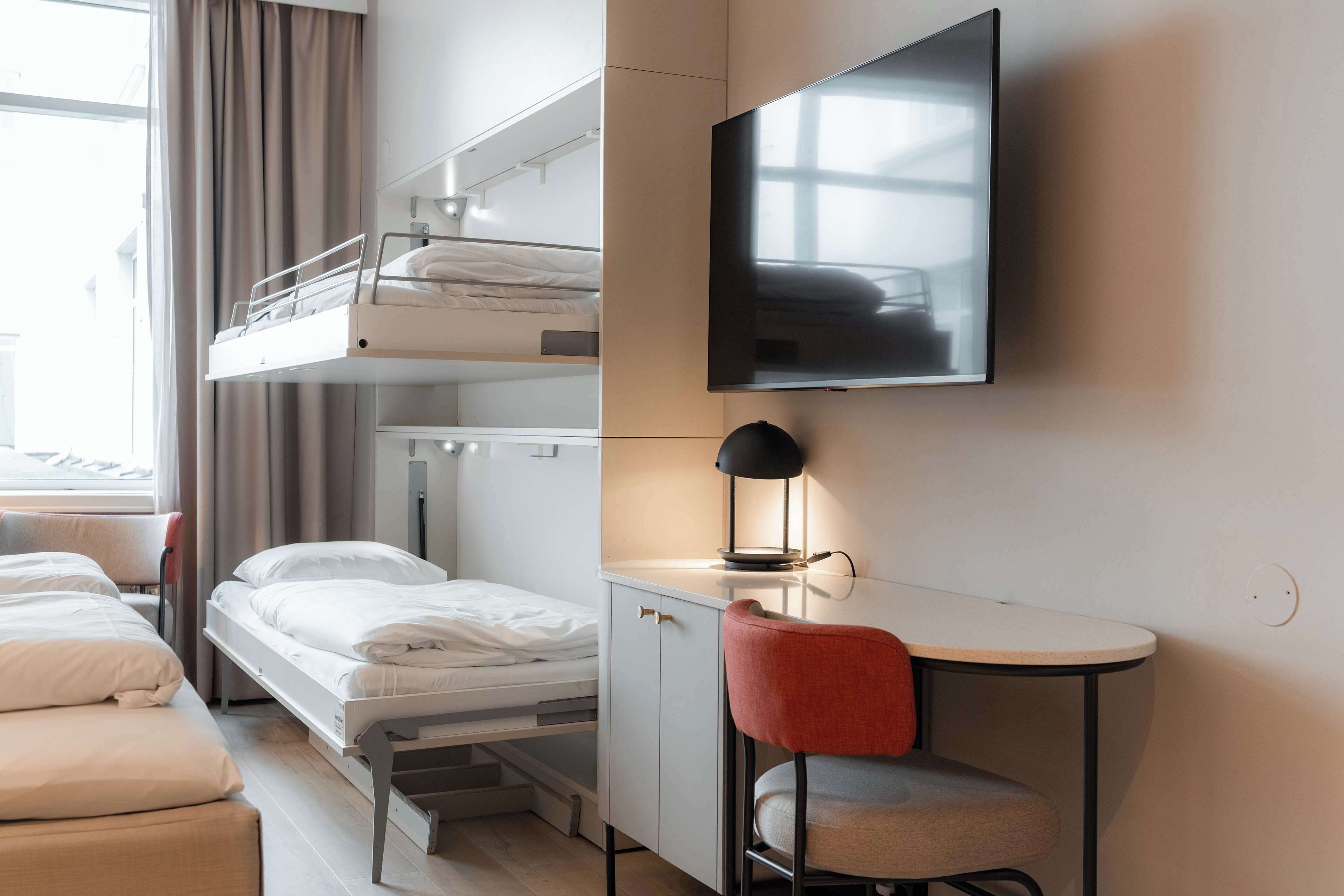 Habitación de hotel con literas, un escritorio, una silla y un televisor montado en la pared.
