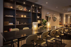 A modern dining area with a long table, multiple chairs, and a bookshelf filled with books and decorative items. - Scandic Oslo City (Oslo)