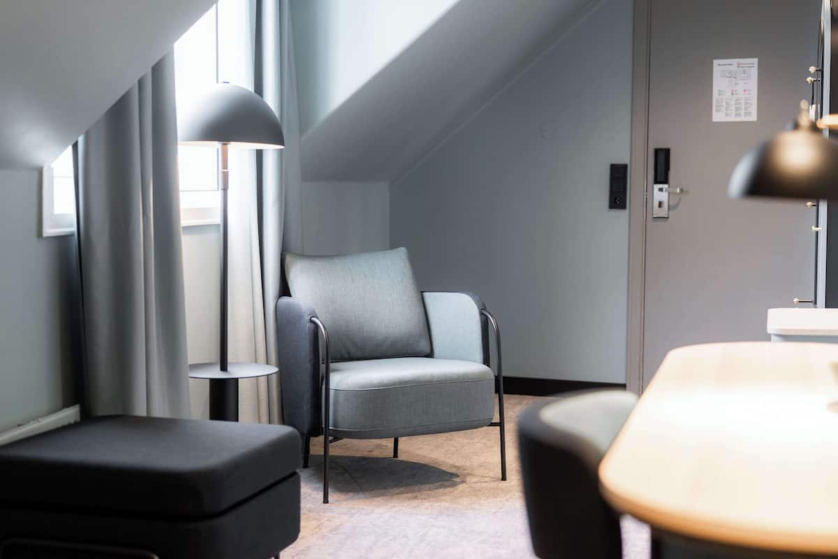 A modern hotel room with a grey upholstered armchair, a black ottoman, and a floor lamp.