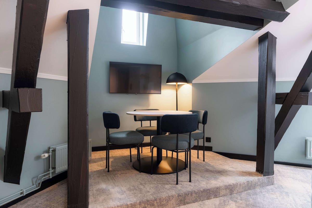 A room with a round table, six chairs, a TV mounted on the wall, and a lamp.