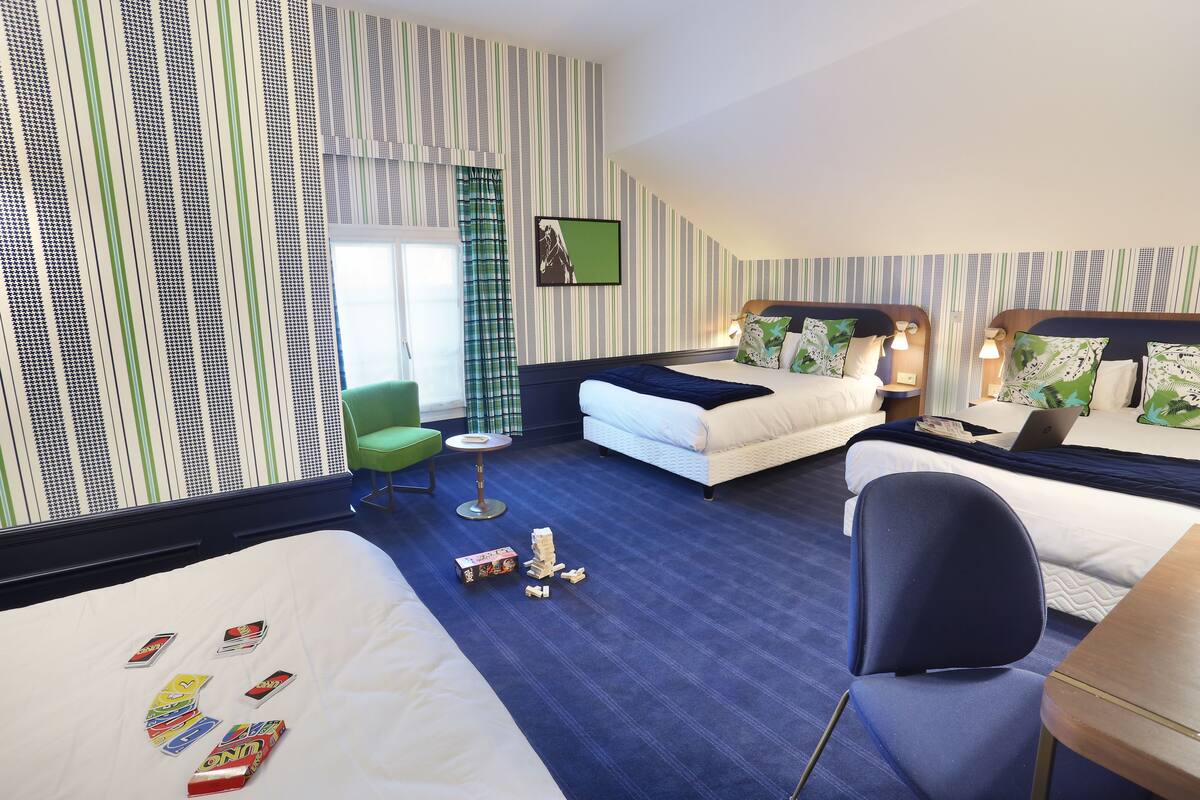 A hotel room with a large bed, a smaller bed, a desk, and a chair. There are striped walls and a window with curtains.