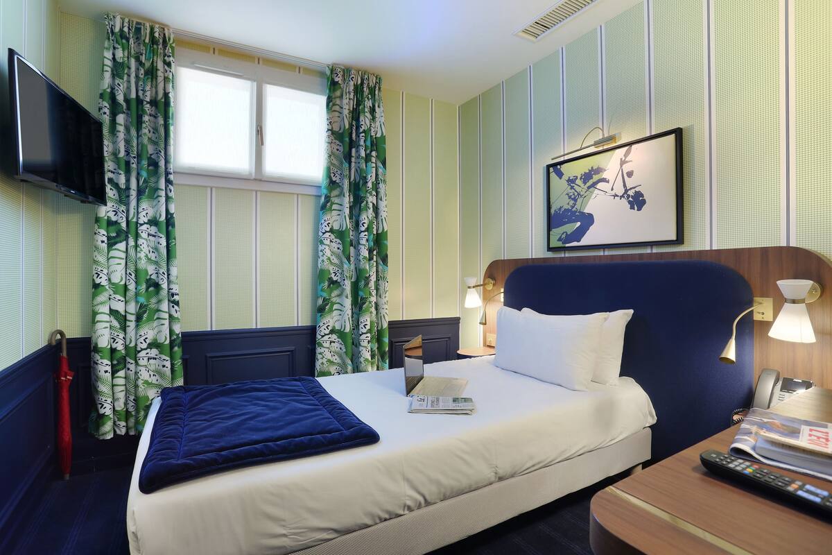 A hotel room with a bed, a desk with a lamp, a TV mounted on the wall, and a window with curtains.
