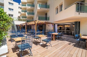 En udendørs terrasse med flere borde og stole, et overdækket siddeområde og udsigt til en bygning med mange etager og altaner.