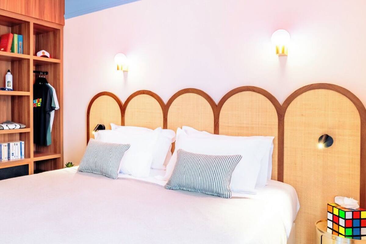 A neatly made bed with a wooden headboard, a bedside table with a Rubik's cube, and a bookshelf with various items.
