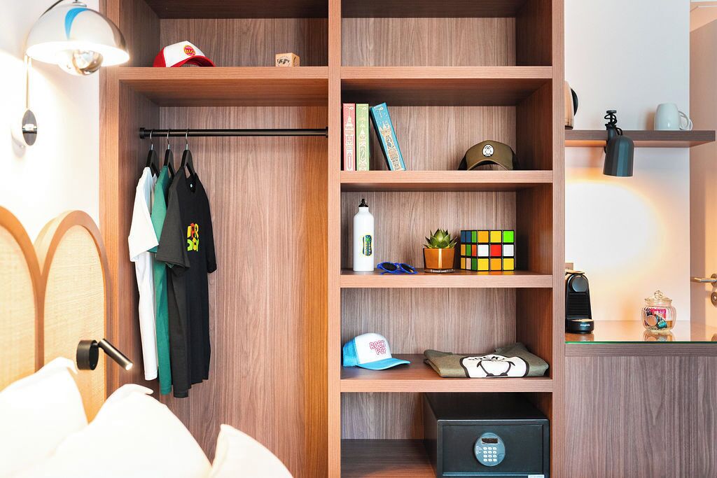 A hotel room with a wooden wardrobe, a bed with white bedding, a small desk with a lamp, and a shelf with books and a Rubik's cube.