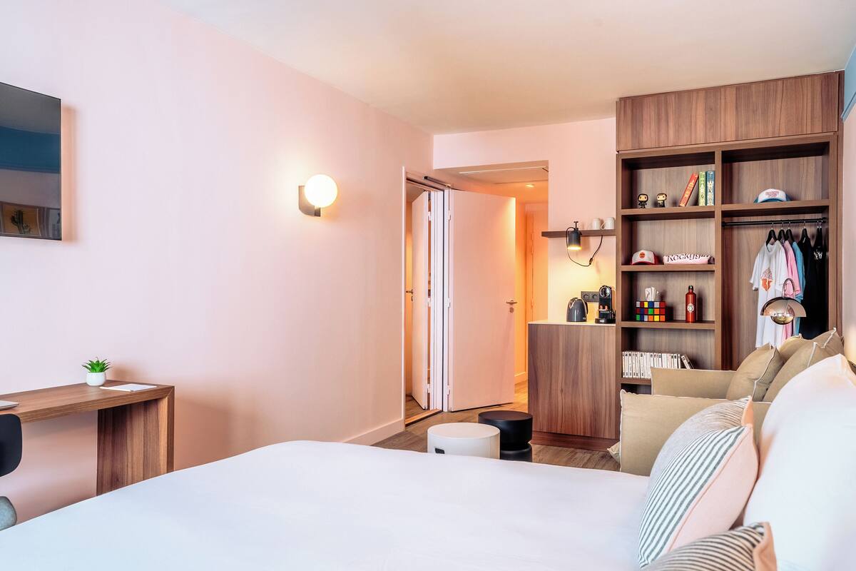A modern hotel room with a bed, a desk, a wardrobe, and a seating area.