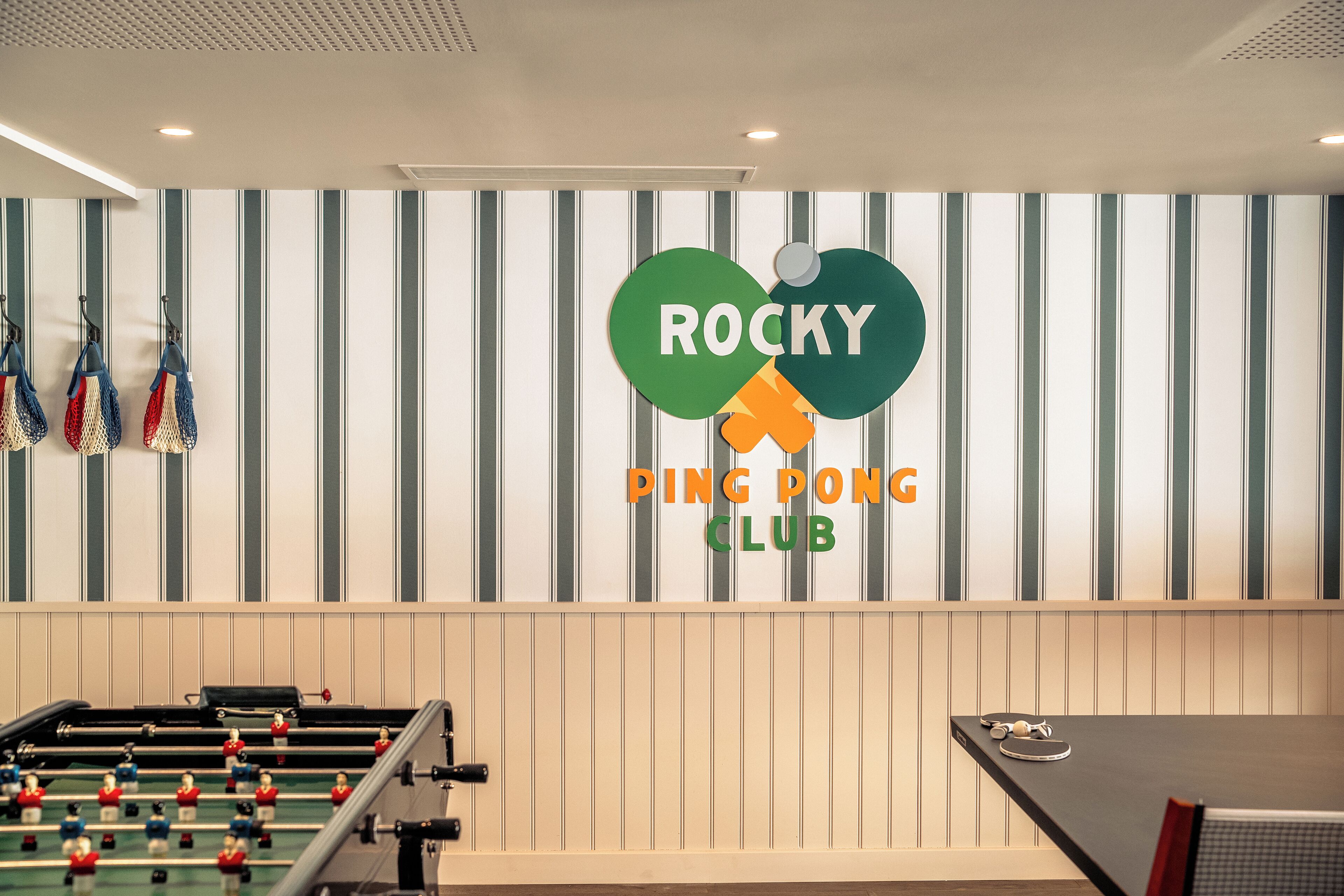 A foosball table and a ping pong table in a room with striped walls and a logo for Rocky Ping Pong Club.