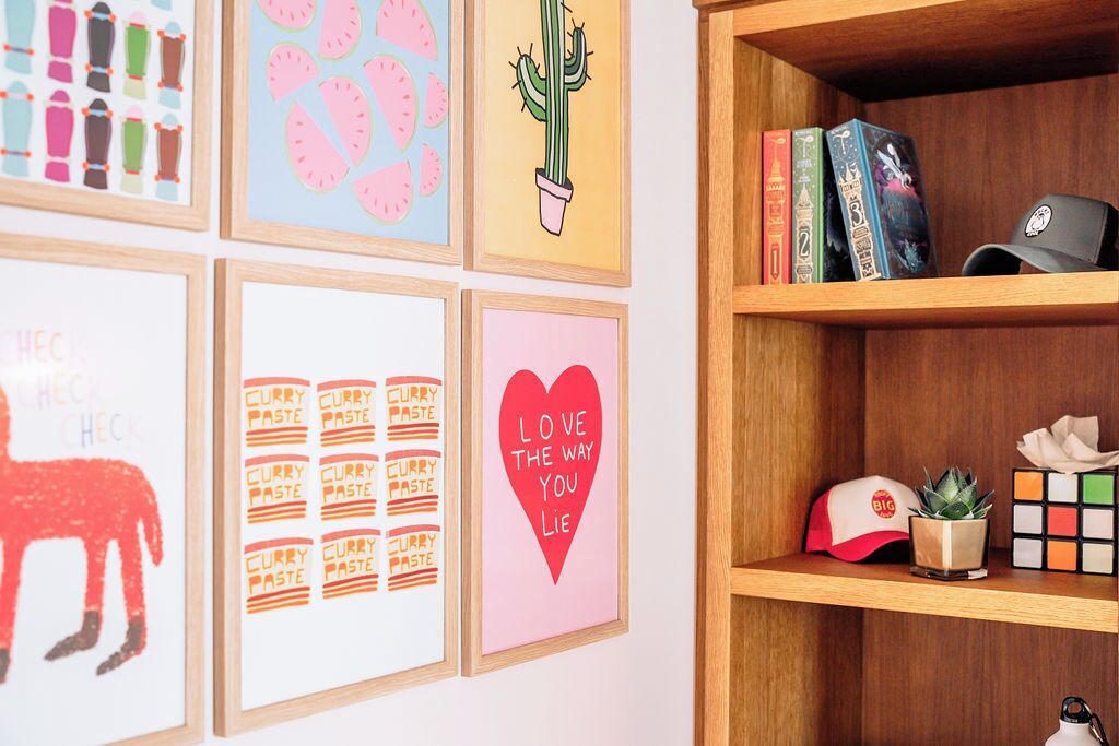 Un mur orné de tableaux encadrés, une bibliothèque remplie de livres et un Rubik’s Cube, et une étagère avec une casquette et une petite plante.