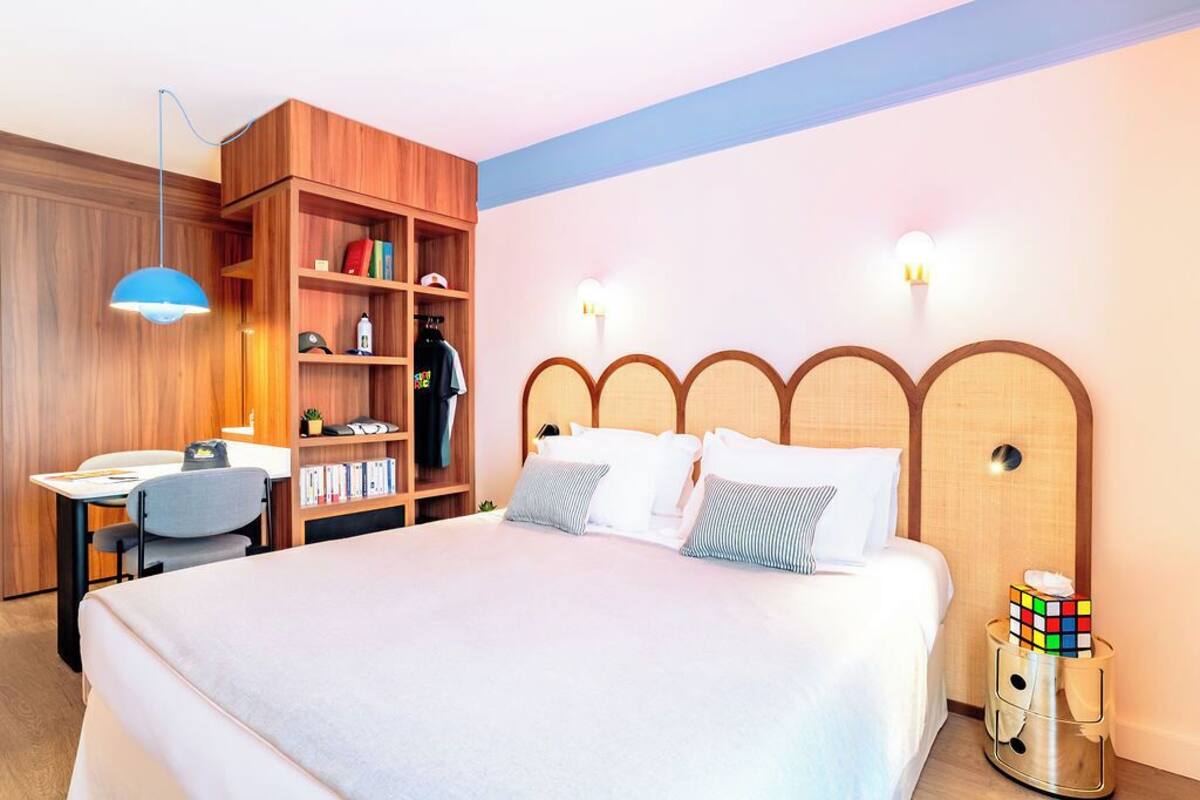 A hotel room with a bed, a desk, a chair, a bookshelf, and a bedside table with a Rubik's cube.