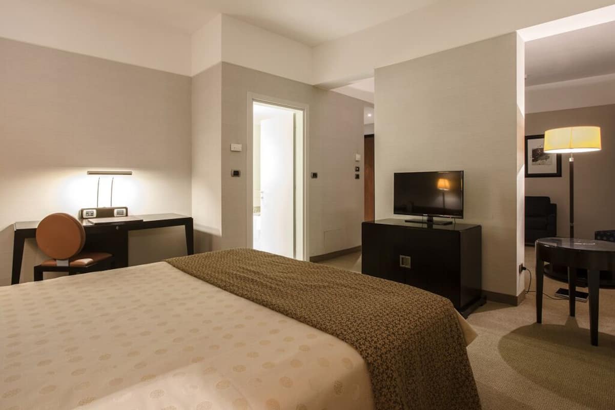 A hotel room with a bed, a desk with a lamp, a television, and a chair.