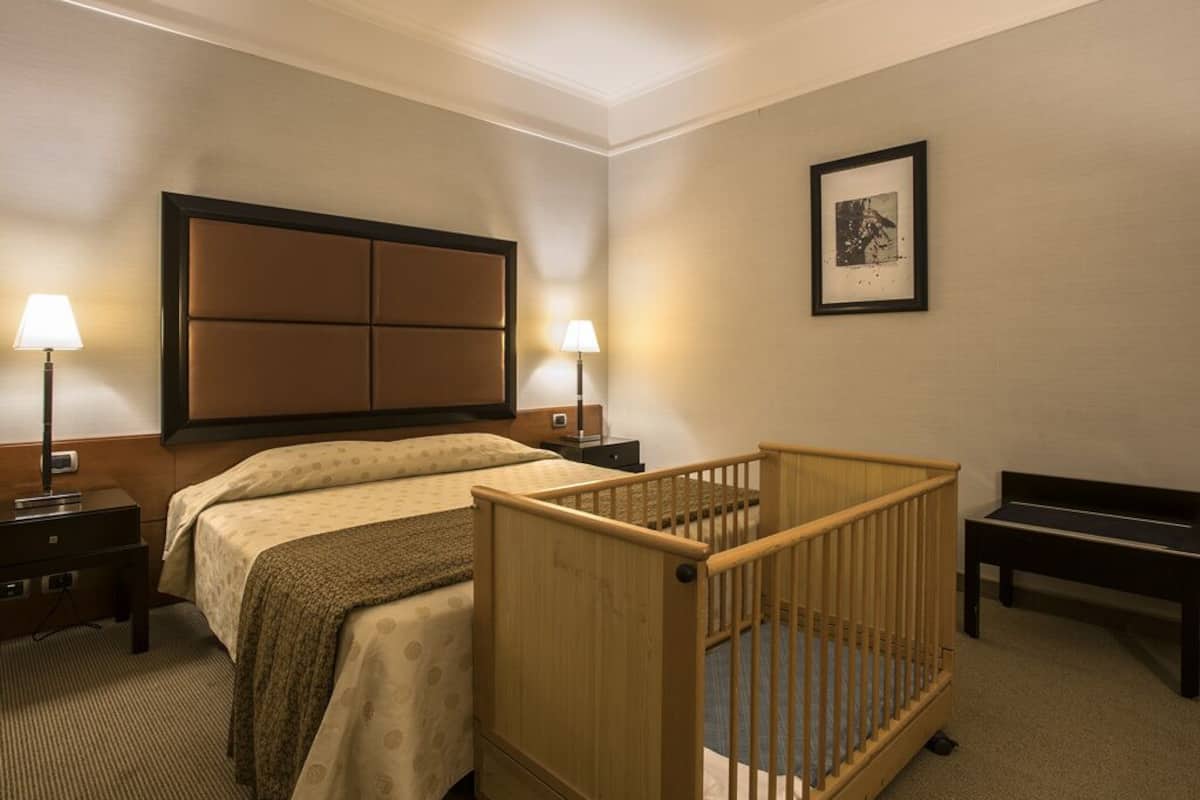 A hotel room with a large bed, two bedside lamps, a crib, a bench, and a framed picture on the wall.