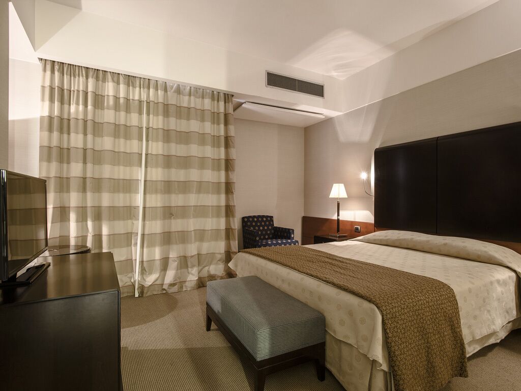 Quarto de hotel com cama, escrivaninha, cadeira, luminária e televisão.