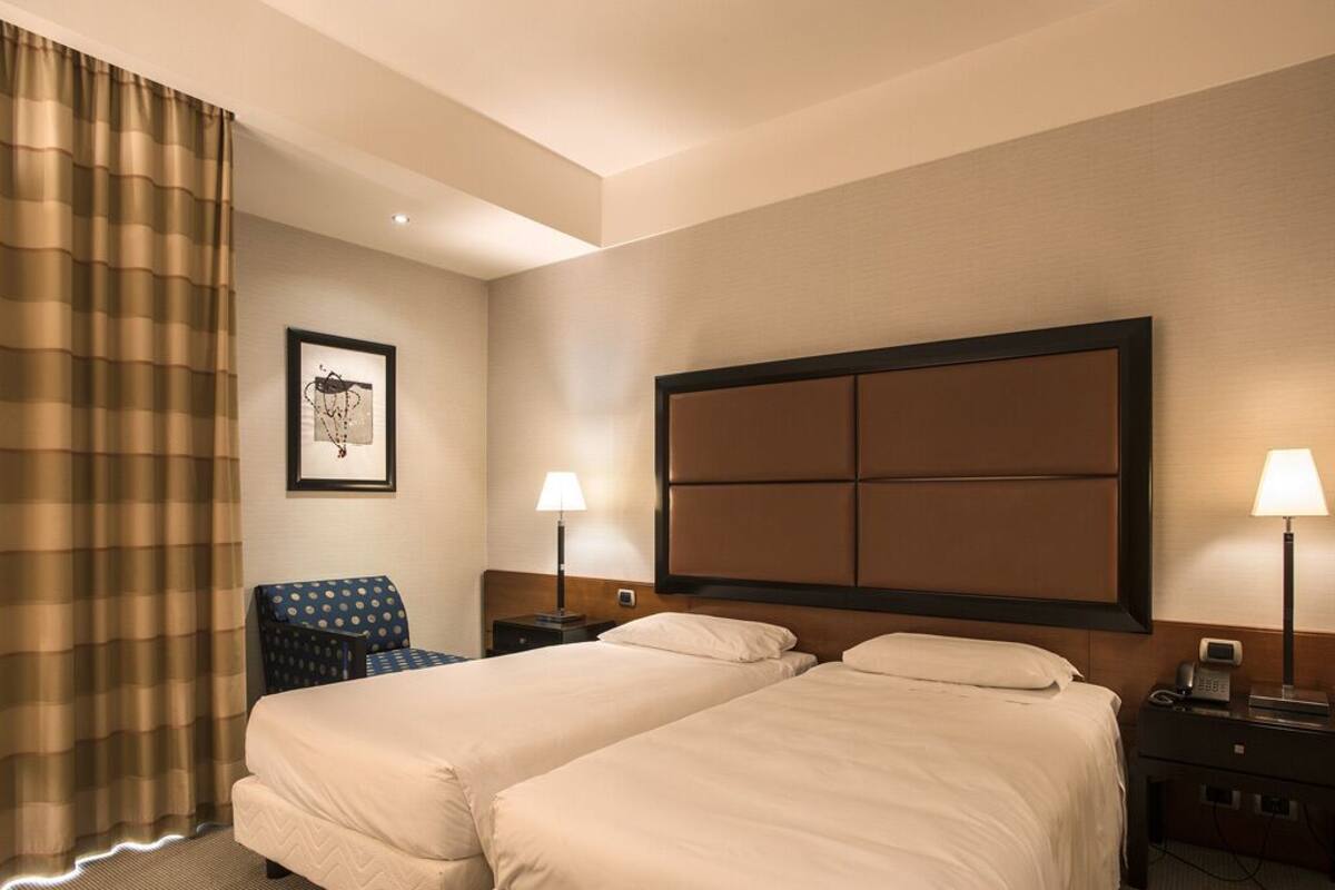 A hotel room with two beds, a chair, a nightstand with a lamp, and a framed picture on the wall.