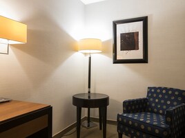 Een hotelkamer met een houten bureau, een rond zwart bijzettafeltje, een vloerlamp, een blauwe fauteuil met patroon en een lijst met abstracte kunst aan de muur.