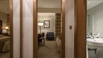 A hotel room with a bed, a desk, a chair, a lamp, a framed picture on the wall, and a bathroom with a sink and mirror.