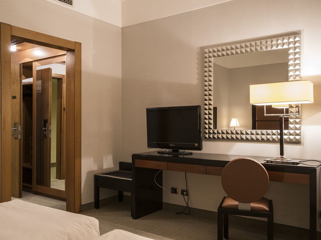 Camera d'albergo con un letto, una scrivania con televisore, una sedia e uno specchio con decorazioni geometriche alle pareti.